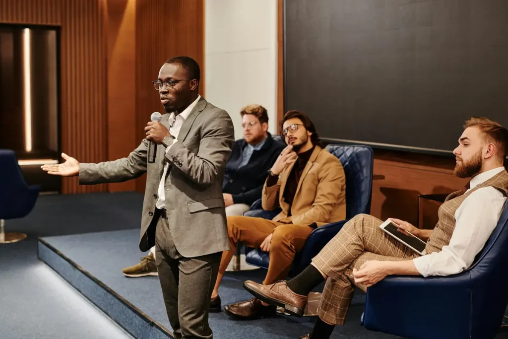 A man is standing to speak at a microphone to ask his coworkers ice breaker questions at a work event. There are three other men sitting behind him, thoughtfully listening. They're all dressed in business attire.